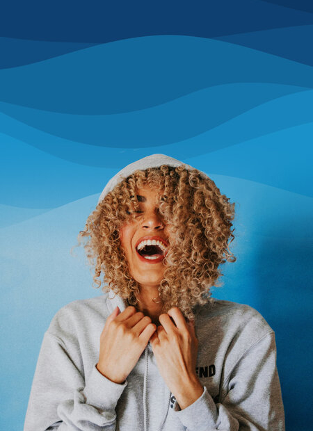 A woman with curly hair laughs while wearing a grey hooded sweatshirt against a blue background